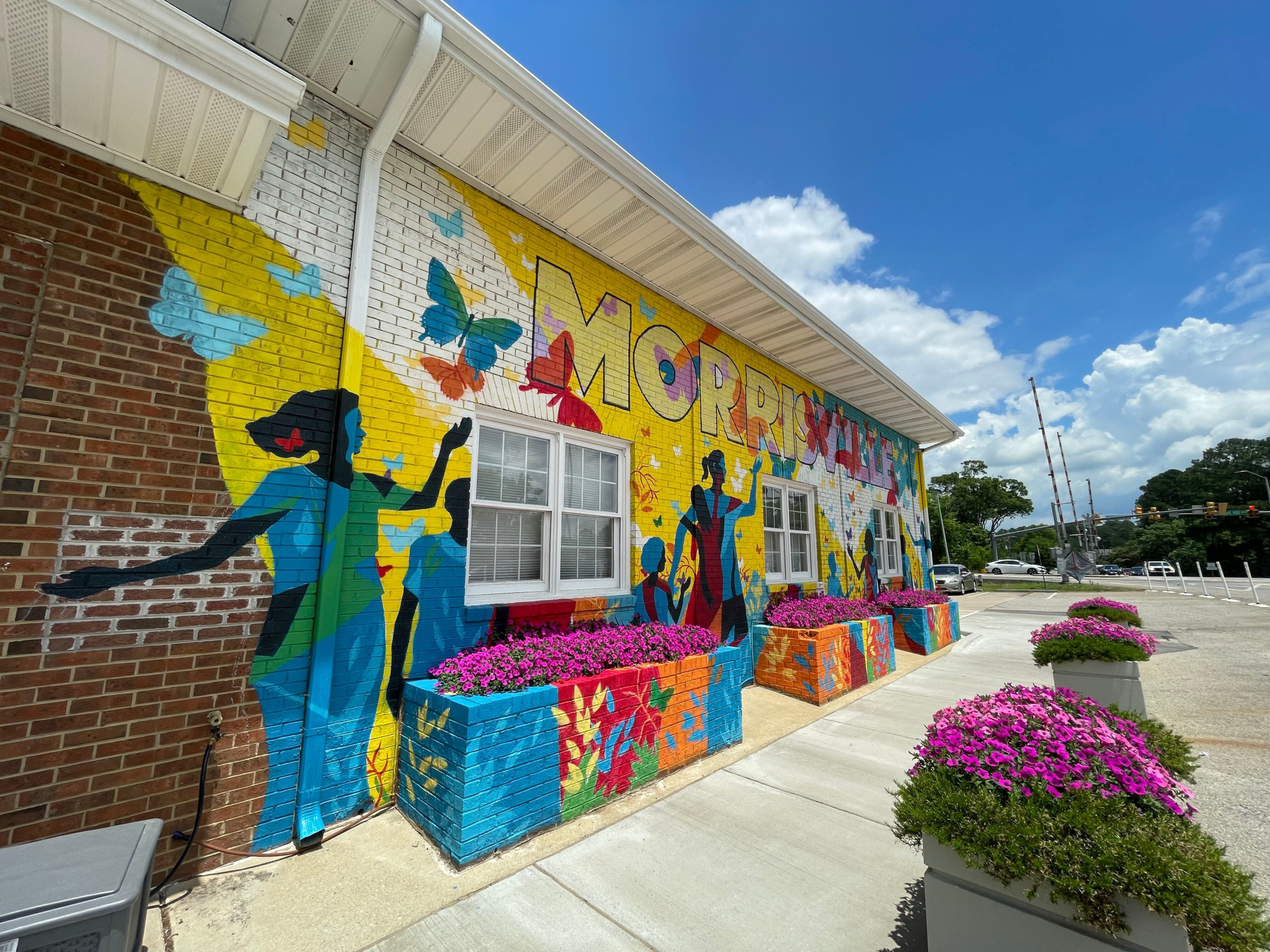 Professional Cleaning Services in Morrisville, NC | Bright & Breezy 1 Iconic public art mural spelling "MORRISVILLE" on a brick building at Shiloh Park in Morrisville, North Carolina, featuring joyful multicultural dancing figures, swirling butterflies, abstract colors, and bright pink flower boxes in the foreground.